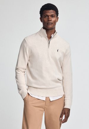 Hellbeige, mit Reißverschluss versehenen Pullover aus weichem Strickmaterial, mit gerippten Bündchen und Saum, und einem kleinen dunklen Logo auf der Brust.