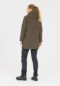 Frau mit blondem Pferdeschwanz, die einen olivgrünen Kapuzenmantel, schwarze Hose und schwarze Stiefel trägt, steht mit dem Rücken zur Kamera vor weißem Hintergrund.