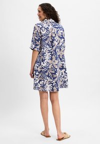 Kurzärmliges, knielanges Kleid mit blauem Hintergrund und weißen sowie beigen Blumenmustern. Der Stoff scheint leicht und lässig geschnitten zu sein.