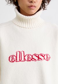 Hvid strikket sweater med en høj ribbet krave. Har et rødt "ellesse" logo i en karakteristisk skrifttype i midten. Blød tekstur.