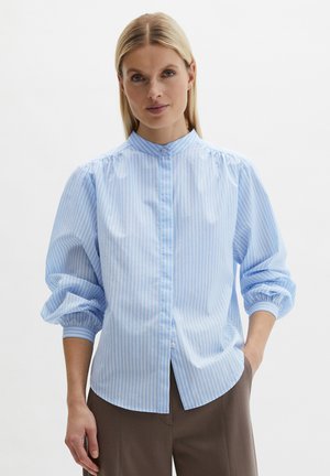 Femme portant une chemise à rayures bleu clair et blanc avec des manches bouffantes et un pantalon marron, sur un fond uni.