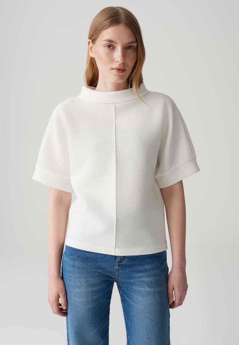 Top bianco a maniche corte con colletto alto, tessuto strutturato e dettagli delle cuciture a contrasto, abbinato a jeans blu.
