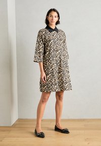 Haljina s leopard uzorkom i crnim ovratnikom, rukavima do tri četvrtine i bočnim džepovima. Model nosi crne ravne cipele na drvenom podu.