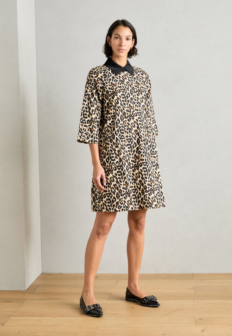 Haljina s leopard uzorkom i crnim ovratnikom, rukavima do tri četvrtine i bočnim džepovima. Model nosi crne ravne cipele na drvenom podu.
