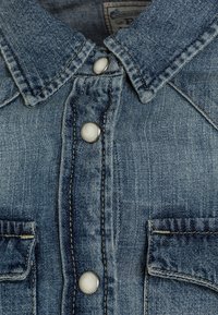 Denimhemd aus hellblauem Stoff, mit klassischem Kragen, silbernen Druckknöpfen und zwei Fronttaschen. Gebrauchter Look und sichtbare Nähte.