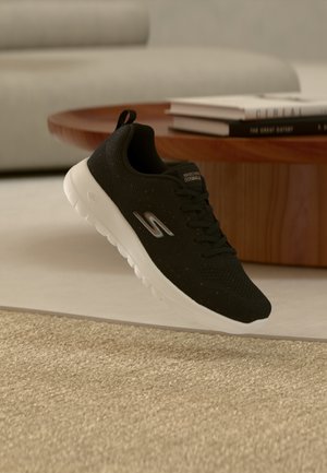 Sort Skechers-sneaker med hvid sål svævende over beige tæppe, rundt træbord og stablede bøger i baggrunden.