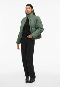 Grøn pufferjakke med høj krave, quiltet design og lynlåslukning, båret over en sort sweater og parret med sorte jeans og hælede støvler.