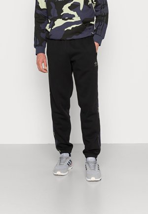 Schwarze Sweatpants in schmaler Passform, mit Seitenstreifen, elastischem Taillengummi und weichem Fleece-Material. Kombiniert mit grauen Sportschuhen.