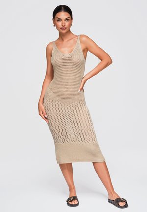 Kvinde poserer i ærmeløs beige strikket midi-kjole med zigzag-mønster og sorte slidesandaler mod en ensfarvet lys baggrund.
