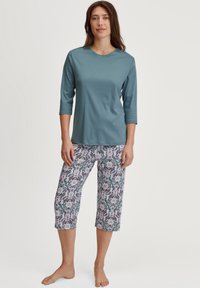 Teal, driekwart mouw top gecombineerd met bloemenpatroon, cropped broek met roze, grijze en zwarte tinten op een lichte achtergrond.