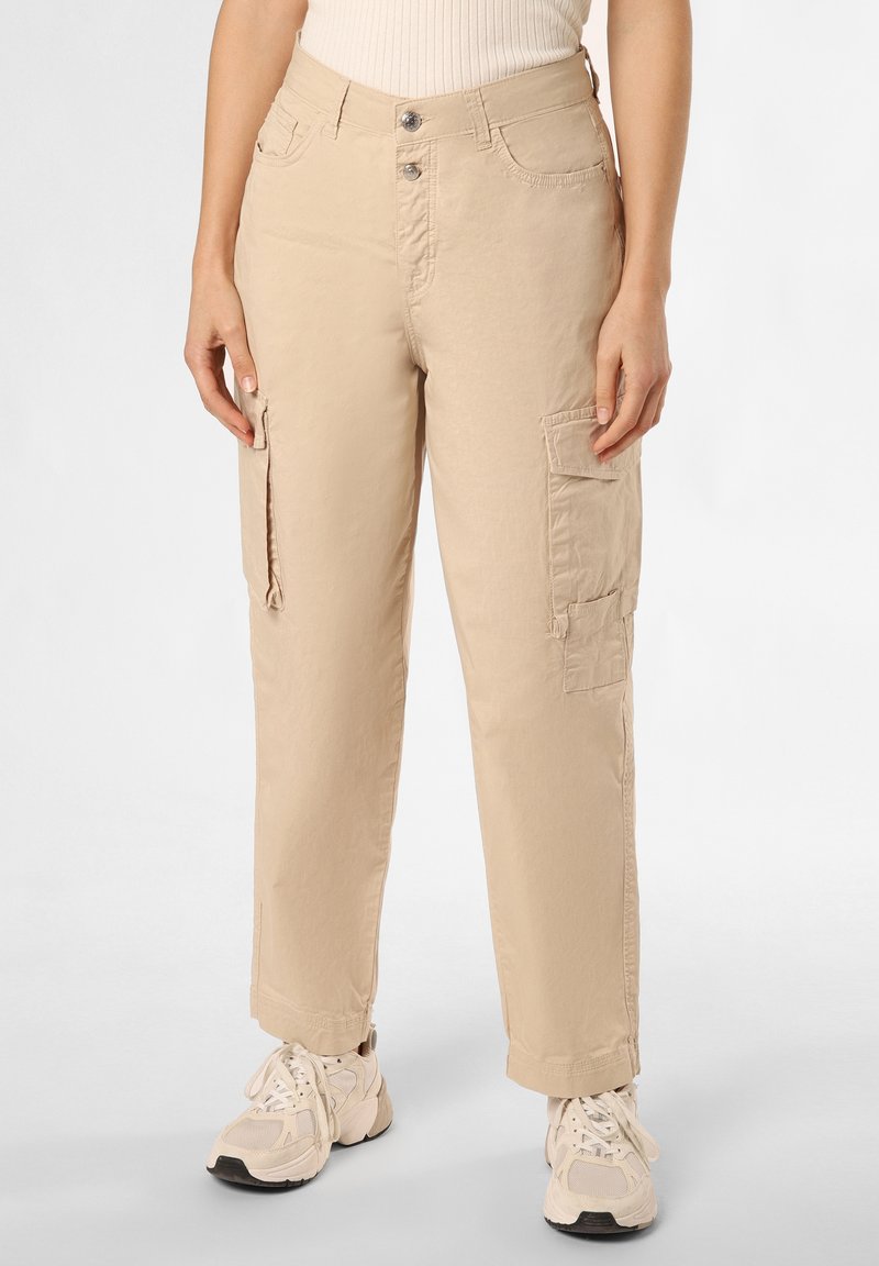 Pantalon cargo beige à multiples poches porté par une personne debout, assorti à des baskets épaisses blanc cassé sur un fond neutre.