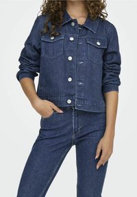 Giacca di jeans blu scuro, con due tasche sul petto, bottoni argentati e orlo arrotondato. Le maniche sono leggermente arricciate ai polsini.