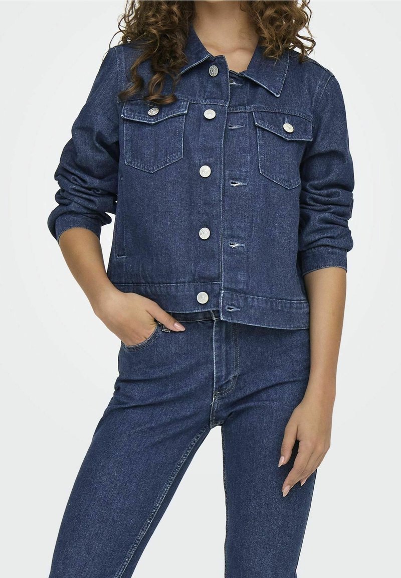 Giacca di jeans blu scuro, con due tasche sul petto, bottoni argentati e orlo arrotondato. Le maniche sono leggermente arricciate ai polsini.