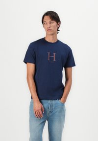Námornícky modré bavlnené tričko s okrúhlym výstrihom, krátkymi rukávmi a hnedým potlačeným logom "H" na hrudi. Kombinované so svetlomodrými džínsami.