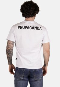 T-shirt bianca in cotone con collo rotondo e maniche corte, caratterizzata da "PROPAGANDA" in grassetto nero sul retro. Design comfort fit.