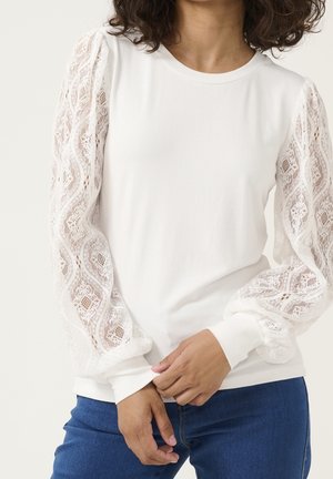 Femme portant un haut blanc à manches longues avec des manches en dentelle et un jean bleu, les mains légèrement croisées au niveau de la taille.