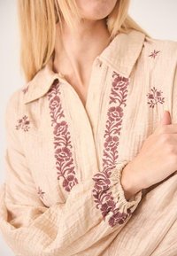 Blusa beige con cuello, con bordado floral burdeos a lo largo de la tapeta y los puños, y detalle de mangas elásticas fruncidas.
