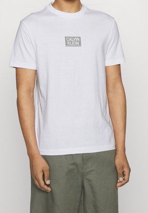 Vit bomullströja med korta ärmar och rundad halsringning. Har en grå rektangulär logotyp med "CALVIN KLEIN" centrerad på bröstet.