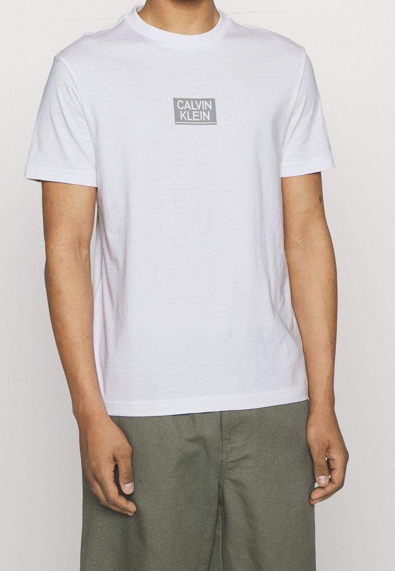 Camiseta de algodón blanca de manga corta y cuello redondo. Presenta un logo rectangular gris con "CALVIN KLEIN" centrado en el pecho.