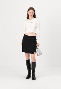 Bijeli rebrasti crop top s crnim logotipom, crna mini suknja s detaljem vezanja sa strane i crne čizme do koljena. Uključuje srebrnu torbicu.