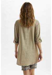 Khaki linnen overhemd met opgerolde mouwen, losse pasvorm en gebogen zoom, gecombineerd met camouflage shorts. Achtergrond is effen wit.