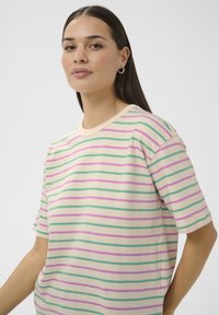 Frau mit langen dunklen Haaren, die Creolen trägt und ein cremefarbenes T-Shirt mit grünen und pinkfarbenen horizontalen Streifen vor einem einfarbigen Hintergrund trägt.