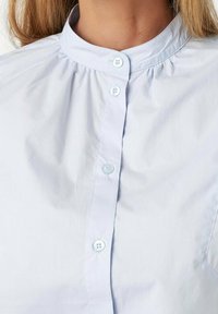 Light blue button-up shirt with a stand collar, featuring a pleated design and small buttons down the front. Smooth fabric texture.