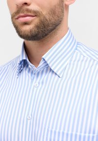Primo piano di un uomo che indossa una camicia button-up a righe verticali blu chiaro e bianche, con colletto a punta e taschino.