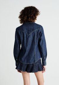 Dark denim shirt with a pointed yoke, vertical seams, and button accents on cuffs, paired with a matching flared denim skirt.