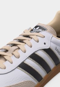 Λευκό sneaker Adidas Samba με μαύρες ρίγες, μπεζ κορδόνια, σουέντ λεπτομέρειες και σόλα από καουτσούκ, που δείχνει το λογότυπο στη γλώσσα και τη σήμανση στο πλευρό.