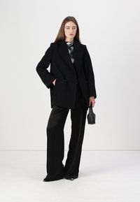 Cappotto doppiopetto nero con ampi rever, abbinato a pantaloni neri a vita alta. Il modello tiene una piccola borsa nera. Sfondo bianco.