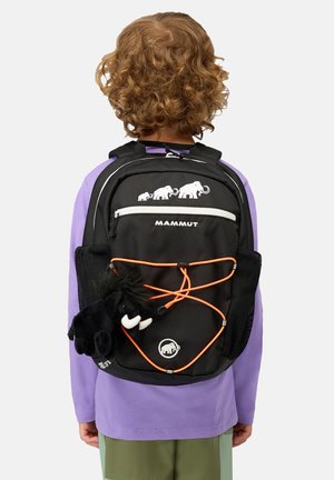 Bambino con capelli ricci che indossa una maglietta viola e pantaloni verdi, che porta uno zaino Mammut nero con lacci arancioni e un peluche mammut nero attaccato.