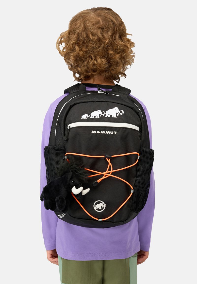 Barn med lockigt hår, klädd i lila tröja och gröna byxor, som bär en svart Mammut-ryggsäck med orange snoddar och en fäst svart mammut-plushleksak.
