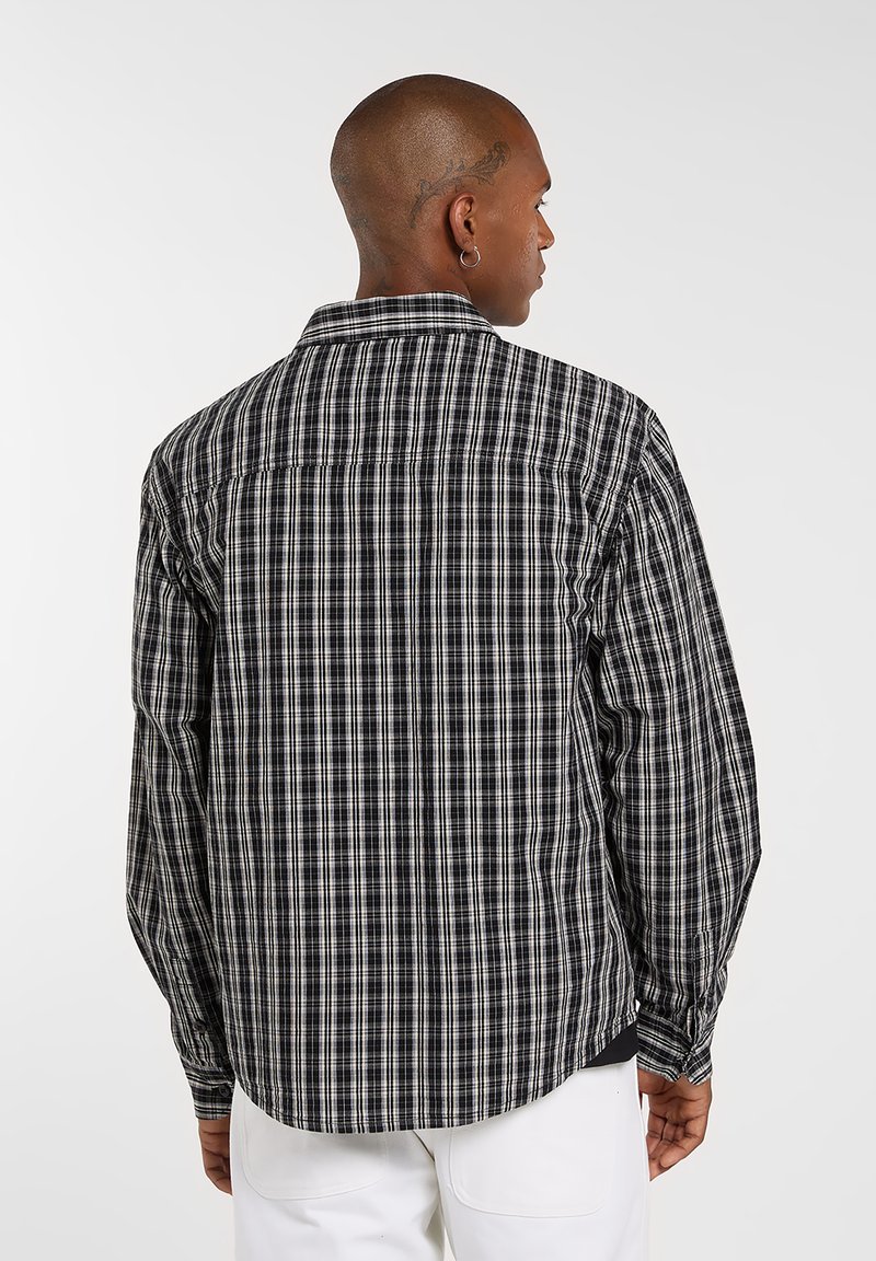 Camicia a quadri bianco e nero, maniche lunghe, colletto con bottoni e orlo curvo. Tessuto in cotone con una consistenza liscia, presenta una piega sul retro.