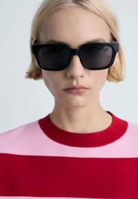 Lunettes de soleil noires rectangulaires avec des verres foncés. Le modèle porte un pull rayé rouge et rose avec un col côtelé. Cheveux blonds courts visibles.