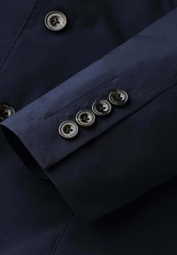 Rukáv námořnického blejzru se čtyřmi tmavými knoflíky z želvovinového vzoru značky Hackett London a detailním prošíváním na hladké látce.