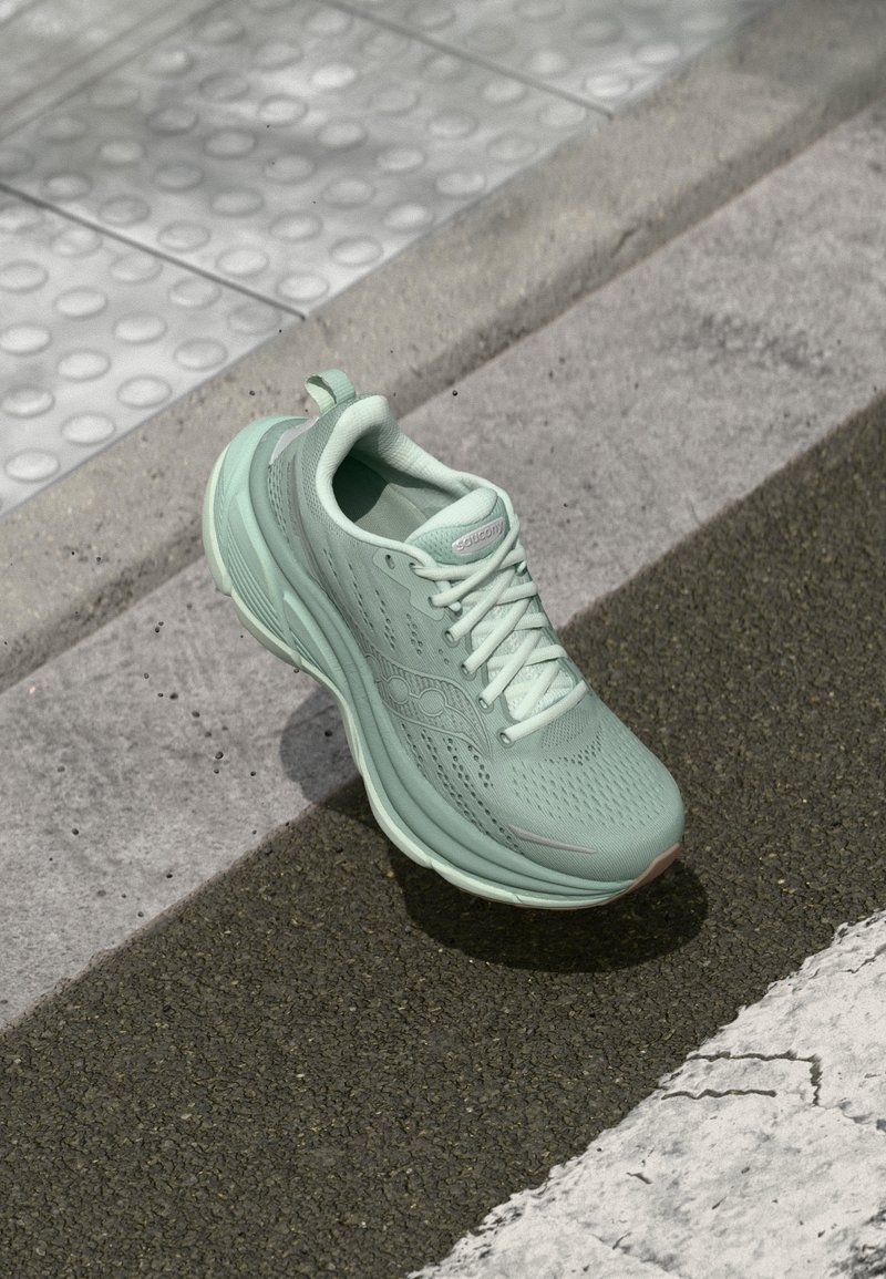 A mint green running shoe with a textured mesh upper, lace-up design, and cushioned sole, positioned on a textured gray sidewalk.