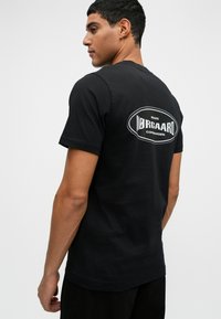 Schwarzes Baumwoll-T-Shirt mit einem runden weißen Logo auf dem Rücken. Das Design ist schlicht mit einem klassischen Rundhalsausschnitt und kurzen Ärmeln.