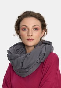 Młoda kobieta o jasnej karnacji, z krótkimi, kręconymi włosami, w dużym szarym szalu i magentowej bluzce, patrząca prosto w obiektyw z neutralnym wyrazem twarzy.