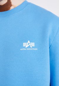 Felpa azzurra chiara realizzata in tessuto misto cotone, con il logo bianco "Alpha Industries" e un design geometrico sul petto.