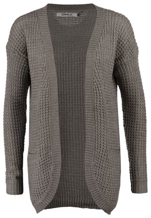 Femme marchant portant un cardigan gris tricoté, un haut bordeaux, un jean bleu, des chaussures blanches et tenant une pochette beige.