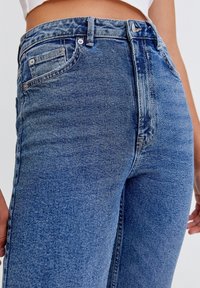 Jeans de denim azul de cintura alta con un acabado ligeramente desgastado, cinco bolsillos y cierre de botón, que presentan un diseño clásico de pierna recta.