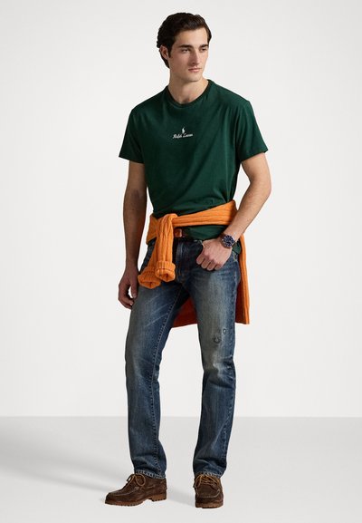 Tricou verde din bumbac cu logo alb, pulover portocaliu legat în jurul taliei, blugi blue denim și pantofi de tip mocasin maro.
