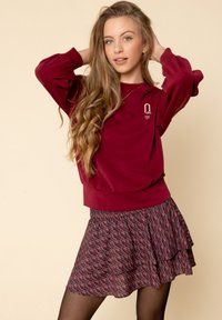 Bordeaux sweatshirt met lange mouwen heeft een geribbelde zoom; gecombineerd met een gelaagde rok met een geometrisch patroon in rood en marineblauw.