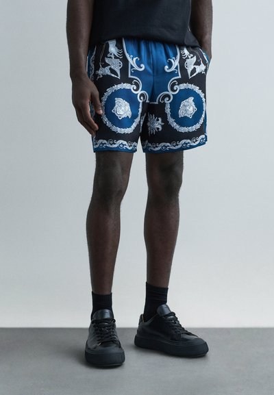 Shorts à motifs noirs et bleus avec taille élastique, présentant des dessins argentés complexes et des motifs de lions. Associés à des baskets noires.