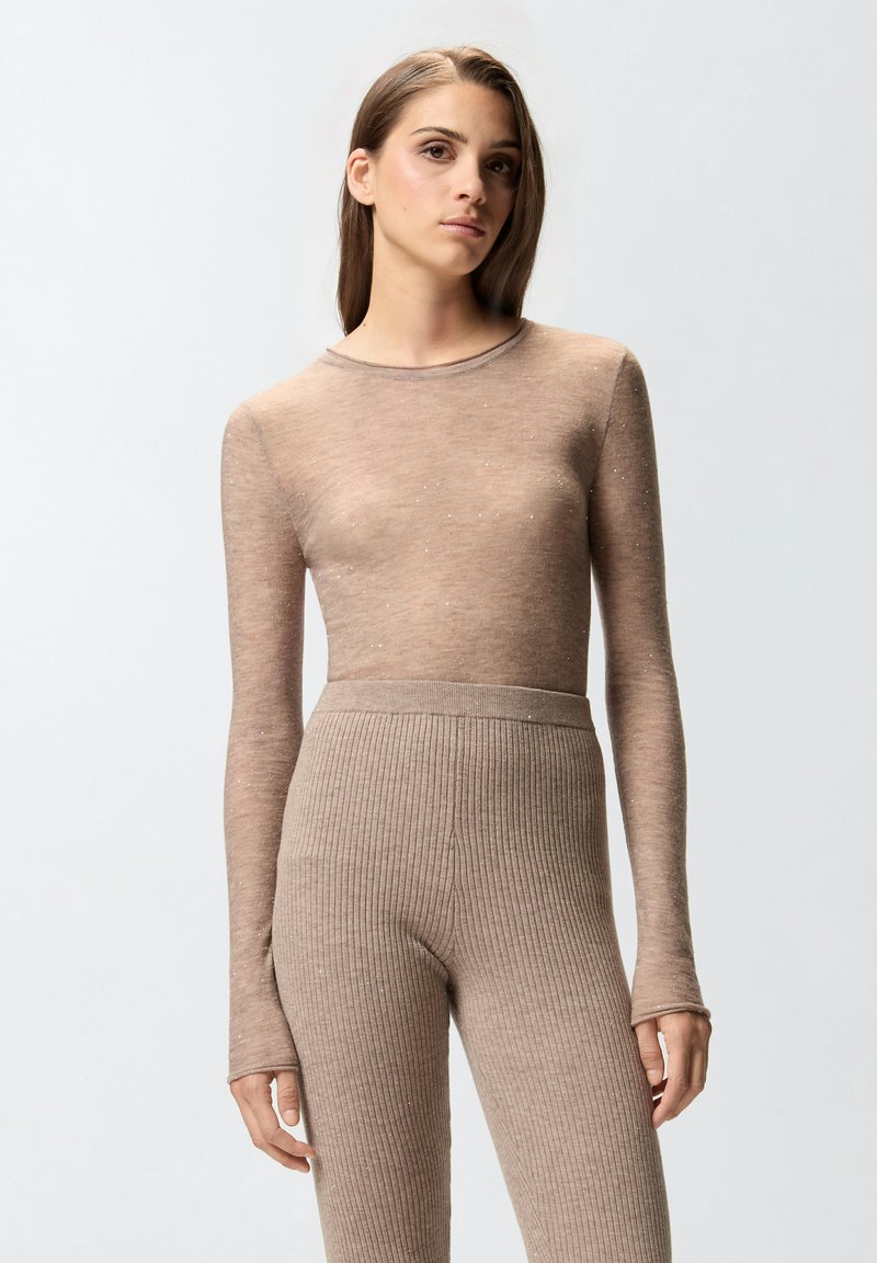 Svijetlosmeđa dukserica dugih rukava napravljena od prozirnog materijala s suptilnim sjajnim detaljima, uparena s rebrastim, visokim svijetlosmeđim tajicama.