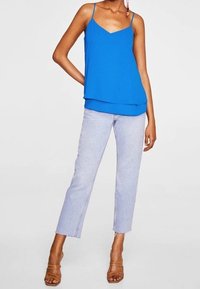 Top sans manches bleu cobalt avec un design superposé, associé à un pantalon cropped en denim clair à coupe droite et coutures visibles. Sandales brunes à lanières.