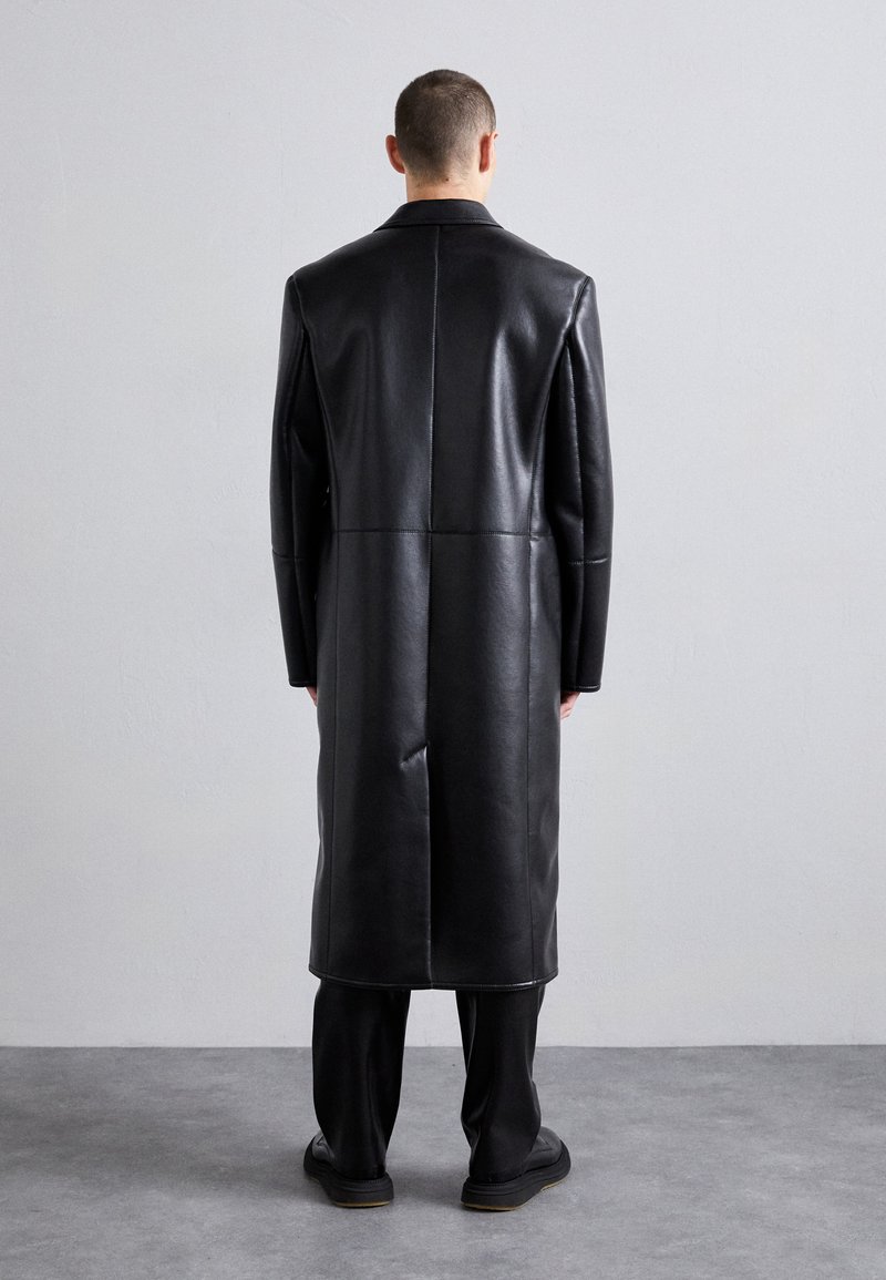 Black leather overcoat with a straight cut, featuring smooth texture and minimal seam detailing. Worn with black trousers and shoes.