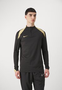 Fekete, hosszú ujjú sportfelső magas nyakkal, arany vállpántokkal és negyed cipzárral. A mellkason egy kis Nike-logo található.