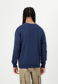 T tumšzila džemperis ar brīvu piegriezumu, ar ribotām aprocēm un apakšmalu, izgatavots no mīkstas auduma, valkāts virs bēšs krāsas cargo biksēm.